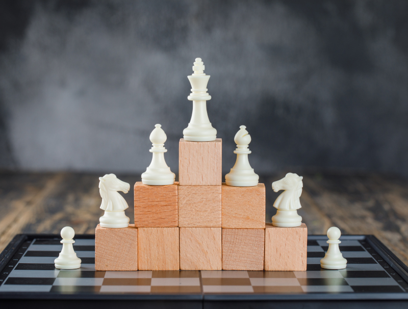 Business hierarchy concept with chessboard, figures on pyramid of wooden blocks on foggy and wooden background side view.